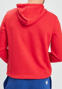 Czerwony hoodie z kapturem, wykonany z miękkiego materiału. Posiada długie rękawy i luźny krój, doskonale komponując się z niebieskimi spodniami. Brak widocznych wzorów.