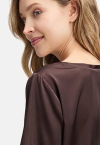 Blouse en satin marron avec une coupe décontractée et une texture douce, dotée d'un col rond et de légères manches bouffantes.