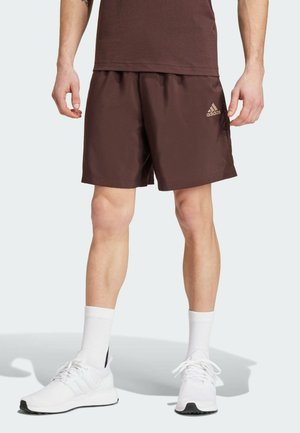 Mężczyzna w brązowych szortach Adidas, białych skarpetkach i białych butach sportowych stoi na jednolitym jasnym tle.