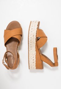 Tan faux leren platform sandalen met gekruiste banden, een geweven jute zool en een gespsluiting rond de enkel. Glad textuur, chunky hak.