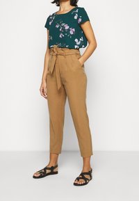 Top floreale verde con maniche corte, abbinato a pantaloni beige a vita alta con cintura a fiocco. Sandali neri piatti con dettagli borchiati.