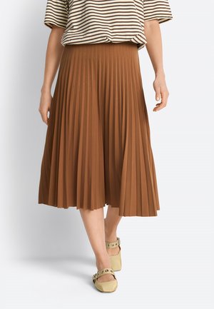 Žena nosi hnedú plisovanú midi sukňu, pruhované tričko s krátkym rukávom a béžové balerínky s remienkami okolo členkov.