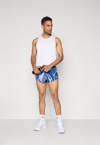 Weißes ärmelloses Tanktop, blaue Shorts mit Wellenmuster, Gewichte haltend, weiße Sneakers und Socken mit schwarzen Akzenten tragend.