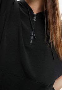 Sudadera negra con cremallera en el frente, cordones y tela suave. Presenta un ajuste relajado y una textura lisa. Acentuada con un diseño de cremallera.