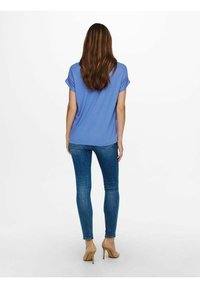 Blå kortermet topp kombinert med tettsittende blå jeans og beige høye hæler, sett fra baksiden. Toppen har rullet ermer og en ledig passform.