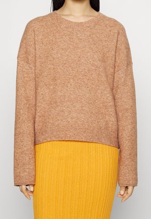 Femme portant un pull en laine brun clair à manches longues et une jupe côtelée jaune, montrée des épaules aux hanches.