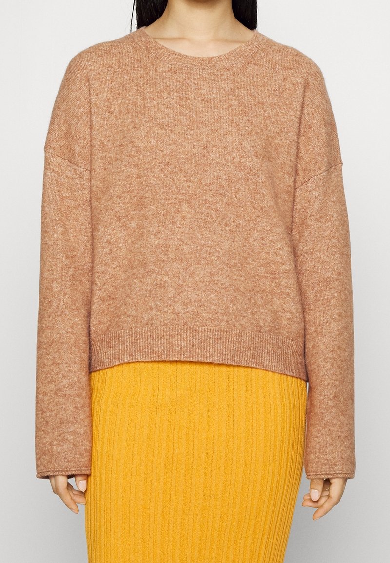 Femme portant un pull en laine brun clair à manches longues et une jupe côtelée jaune, montrée des épaules aux hanches.