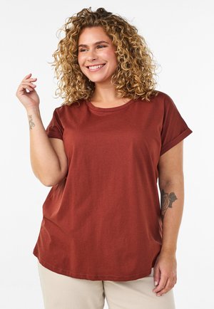 Smilende kvinde med krøllet blondt hår iført en rustfarvet t-shirt og beige bukser, der viser tatoveringer på begge arme.