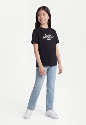 Jeune fille souriante aux longs cheveux bruns portant un t-shirt noir avec un texte blanc, un jean bleu clair et des baskets blanches, debout sur un fond blanc.