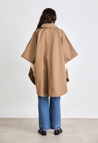 Poncho en coton marron, avec un large col, des manches amples et une coupe fluide, associé à un jean bleu taille haute et des chaussures noires.