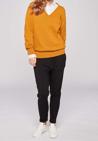 Gul v-ringad tröja med ribbad textur, kombinerad med svarta smala byxor och vita sneakers. Synligt varumärkeslogo på tröjan.