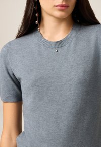 Lyseblå kortærmet sweater af blødt stof med ribbet halsudskæring og en lille dekorativ detalje foran.