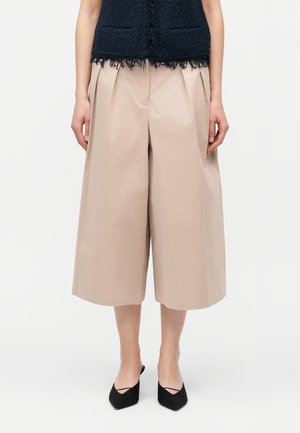 Femeie care poartă culottes bej cu picioare largi, top negru fără mâneci cu tiv cu franjuri și pantofi negri cu vârf ascuțit pe fundal alb.