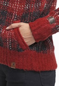 Suéter de punto en rojo y negro, con un diseño texturizado, puños acanalados y una etiqueta de cuero en el dobladillo. Bolsillo frontal visible.