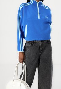 Pullover in pile blu con colletto con zip, caratterizzato da righe azzurre sulle maniche e tasche laterali. Abbinato a jeans neri e una borsa bianca.