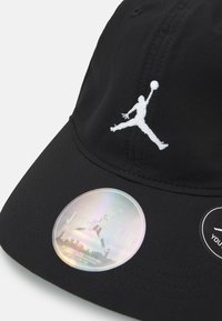 Căciulă din țesătură neagră, cu logo-ul Jumpman brodat în alb. Include un autocolant holografic cu textul "JORDAN" și un tag circular negru suplimentar.