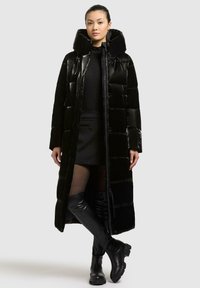 Cappotto trapuntato nero con collo alto, texture lucida, zipper a tutta lunghezza e bottoni a scatto, indossato sopra un outfit aderente e stivali neri fino al ginocchio.