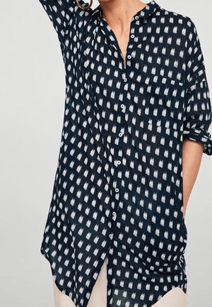 Persona che indossa una camicia blu navy con bottoni, motivi bianchi a forma di diamante, maniche arrotolate e grandi orecchini a cerchio dorati.