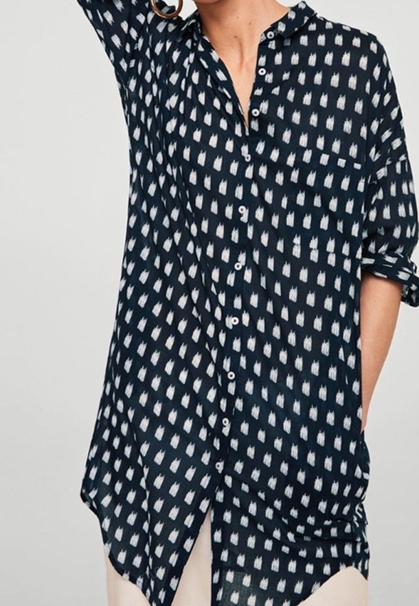 Personne portant une chemise bleu marine à boutons avec des motifs blancs en forme de losange, manches retroussées et grandes boucles d’oreilles créoles dorées.