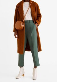 Bruine wollen jas, witte blouse, groene op maat gemaakte broek, witte enkellaarsjes met ritsen en een bruine ronde schoudertas.