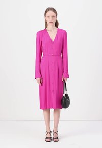 Fuksia pitkähiha dressi, jossa on V-pääntie, nappi edessä ja solmittava vyötärö. Mallilla on musta käsilaukku ja hänellä on päällä mustat halkiot.