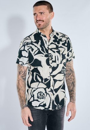 Mann mit kurzen Haaren und Tattoos auf beiden Armen, der ein schwarz-weißes, geblümtes Kurzarmhemd trägt und zur Seite schaut.