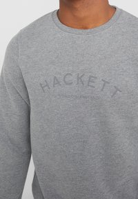 Sudadera gris hecha de material suave, con cuello redondo y un logo desgastado "HACKETT LONDON ENGLAND" en un diseño curvado.