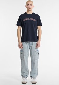 Mornarska kratka majica s kratkim rukavima s logotipom "GUESS JEANS", uparen s svijetloplavi cargo trapericama koje imaju više džepova i opušteni kroj.
