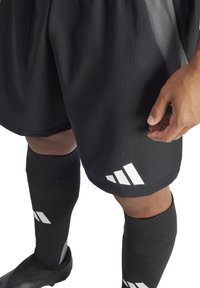 Schwarze Sportshorts mit vertikalen Streifen, die ein weißes Adidas-Logo zeigen. Schwarze Kniesocken vervollständigen das Outfit. Die Struktur des Stoffes ist sichtbar.