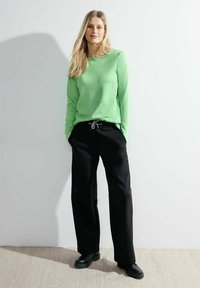 Zelený dlouhý rukáv s texturovaným povrchem, zkombinovaný s černými širokými kalhotami s tkaničkovým pasem. Černá obuv dotváří celkový vzhled.