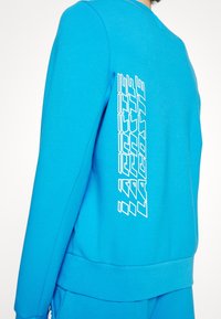 Light blue sweatshirt featuring a ribbed neckline and cuffs. The white, stylized logo is printed vertically on the left side.