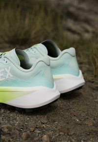 Paire de chaussures de course bleu clair et vert reposant sur un terrain rocheux en plein air, vue de dos pour montrer les détails du talon et de la semelle.