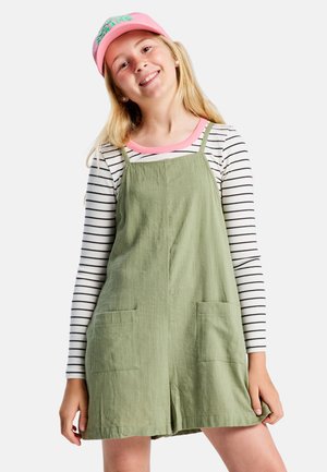 Vestido verde de algodón tipo peto con dos bolsillos delanteros, llevado sobre una camiseta de manga larga a rayas blancas y negras, combinado con una gorra rosa.