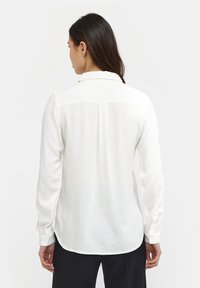 Camicia bianca a maniche lunghe con colletto, leggermente lucida e con un orlo curvo. Presenta pieghe sul retro e polsini con bottoni. Tessuto dalla texture liscia.