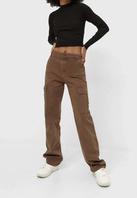 Pantaloni cargo marroni con gamba dritta, dotati di due tasche laterali e dettagli cuciti, abbinati a un top corto a costine nero e sneakers bianche.
