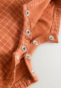 Forbrændt orange onesie stof med et gridmønster, der har sølvknaplukninger langs halsudskæringen og blødekanter.