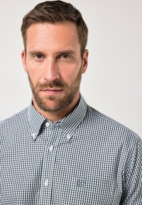Camisa de cuadros en blanco y negro, de botones, con cuello vuelto, que presenta botones blancos y un bolsillo en el pecho izquierdo con un logo.