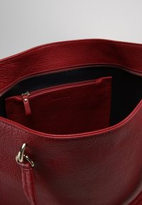 Czerwona skórzana torba typu tote z teksturowaną powierzchnią, szerokim otworem i czarną kieszenią na zamek w środku dla lepszej organizacji. Akcenty w kolorze złotym.