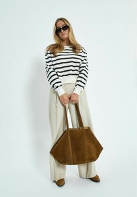 Pružitasti džemper u crno-bijelim tonovima, svijetlosmeđi hlače, smeđa suedna torba i smeđa obuća. Manekenka pozira na svijetloj pozadini.