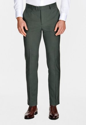 Mężczyzna w ciemnozielonych dopasowanych eleganckich spodniach, białej eleganckiej koszuli oraz wypolerowanych brązowych skórzanych butach, stojący z rękami swobodnie opuszczonymi wzdłuż ciała.