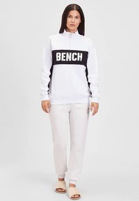 Witte sweatshirt met een zwart paneel waarop het woord "BENCH" staat, ritskraag en lange mouwen, gecombineerd met lichtgrijze joggingbroek en beige slippers.
