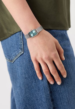 Personne portant une montre à bracelet en maille argentée avec un cadran turquoise carré, main reposant sur la poche d'un jean bleu en denim, chemise vert olive visible.