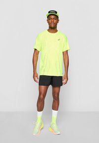 Neon dzeltena sporta krekls ar sieta dizainu, melni šorti, neon zaļas zeķes un pelēki skriešanas apavi ar oranžu zoli. Melna cepure.