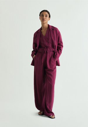 Mujer de pie con las manos en los bolsillos, vistiendo un traje de rayas de ajuste holgado en color magenta oscuro y sandalias negras abiertas en los dedos, sobre un fondo liso.