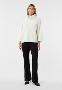 Kobieta ubrana w białą żebrowaną sweter z golfem, czarne spodnie proste i czarne czółenka na szpilce, stojąca na jednolitym tle.