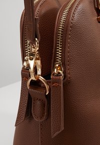 Brown textured leather bag featuring a gold zipper, gold hardware, and a decorative strap detail. Close-up view of the zipper and accents.