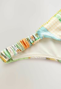 Több színben pompázó bikini felsőrész, amely ráncolt pánttal rendelkezik, zöld, kék és narancssárga mintákkal. Simítás, puha tapintás és sárga díszítő szegély jellemzi.