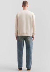 Lichtbeige sweatshirt met geribde manchetten en zoom, gecombineerd met losvallende vervaagde blauwe jeans en zwarte sneakers, gezien vanaf de achterkant.