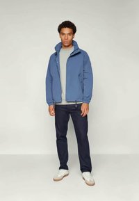 Blue waterproof jacket with a high collar, zip closure, and elastic cuffs, paired with a light gray shirt and dark navy pants.