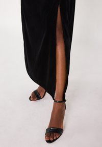 Robe en velours noir avec une fente sur le côté et une longueur aux chevilles. Accompagnée de sandales noires à lanières. Texture lisse avec un léger éclat.